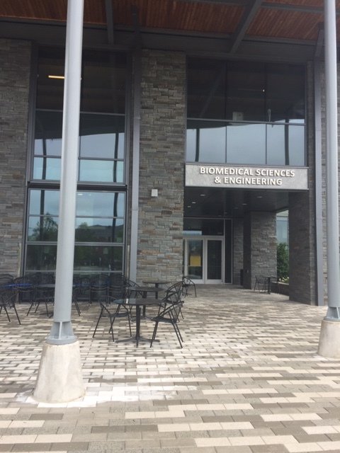 A gray stone building surrounded by a gray brick walkway, black chairs, and round, black tables. The entry reads, "Biomedical Sciences and Engineering."