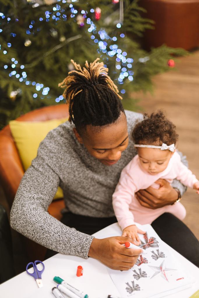 A Black man in a gray sweater coloring in pictures. He is holding his baby daughter, who is wearing pink and a tiny hairbow.