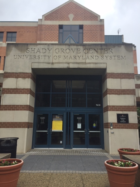 A building entryway with a blue door and horizontal stripes made of brick. Image Text: "Shady Grove Center, University of Maryland System."